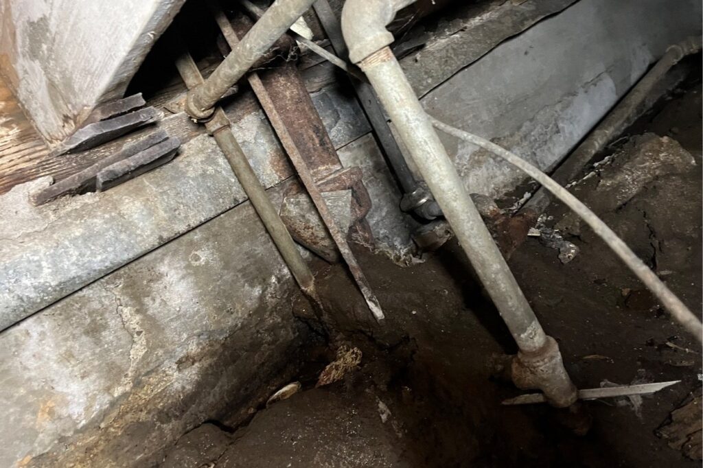 Old galvanized water pipes running down into a crawlspace beside the foundation.