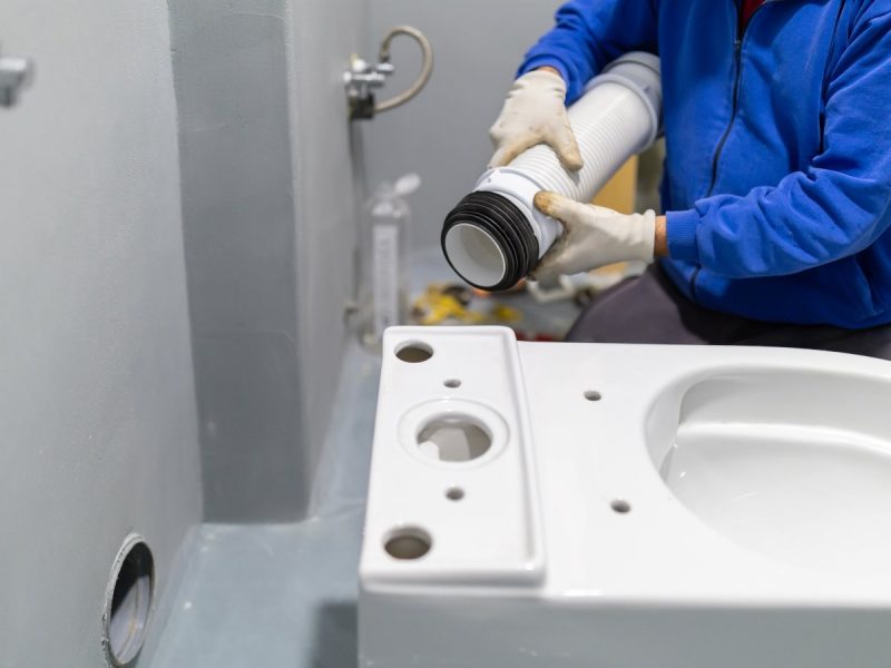 AdobeStock_1769865083 Plumber hands in protective gloves are meticulously connecting a flexible pipe to the rear of a white ceramic toilet bowl, performing a new bathroom plumbing installation. Builders apartment improvement or restoration works